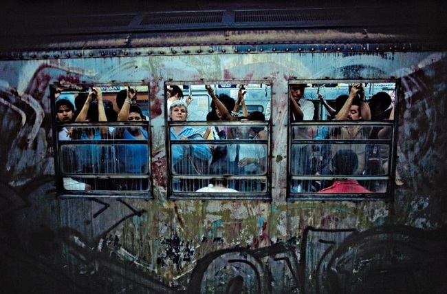 This picture shows a moving train with all kinds of graffiti over it. People are shown travelling in the train. This picture shows a moving train with all kinds of graffiti over it. People are shown travelling in the train.